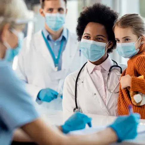 Nurse wearing a mask holding a child while looking toward other medical personnel.